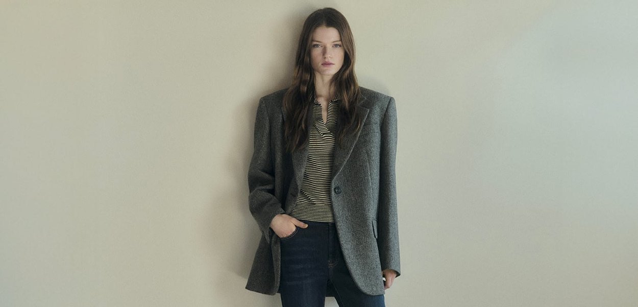 Blazer in misto lana grigia con spalle strutturate, chiusura con un bottone, abbinato a una camicia a righe e jeans scuri, su uno sfondo chiaro.
