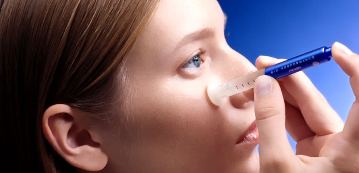 Ein klarer, transparenter Applikator wird in der Nähe des Auges verwendet, gehalten in einer Hand mit einem blauen, stiftähnlichen Gerät, vor einem sanften, verschwommenen blauen Hintergrund.