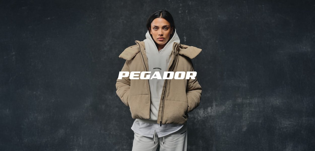 Frau mit dunklem Haar trägt eine beige Steppjacke über einem weißen Hoodie und Hosen, steht vor einem dunklen, strukturierten Hintergrund mit dem Text "PEGADOR".
