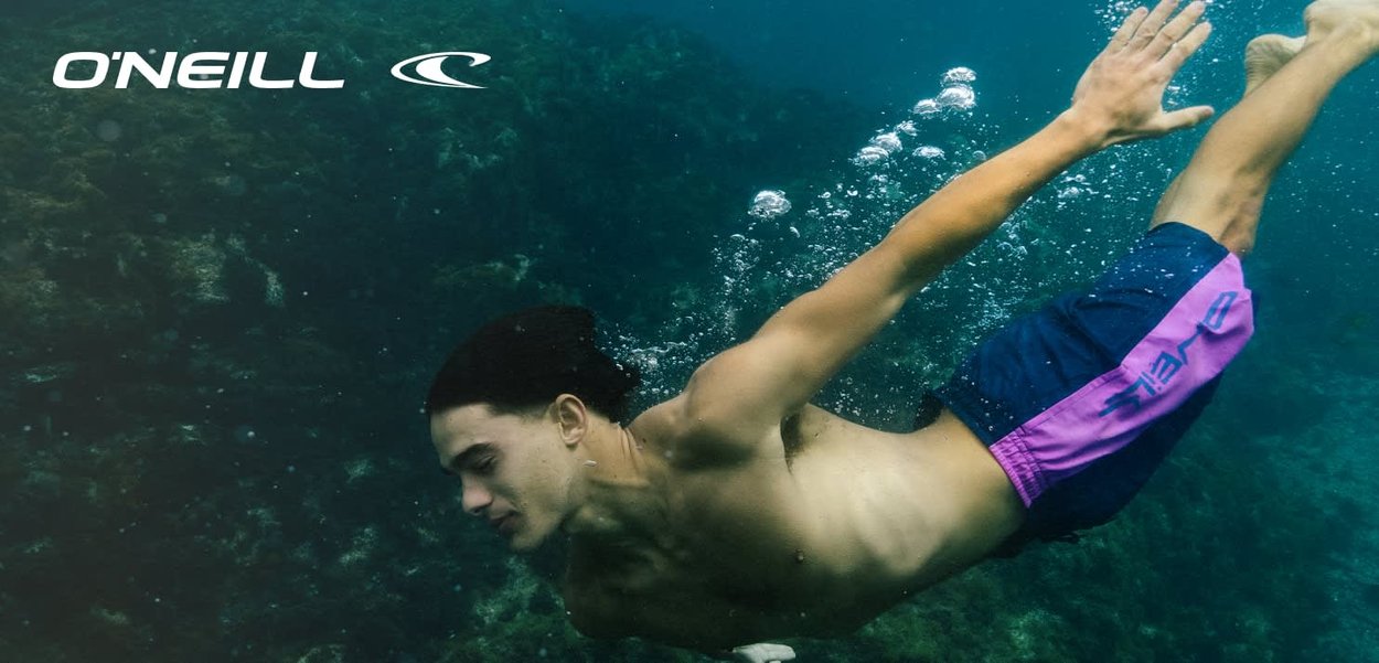 Junger Mann schwimmt unter Wasser in der Nähe eines Korallenriffs und trägt blaue und pinke O'Neill-Badeshorts mit O'Neill-Logo oben links.