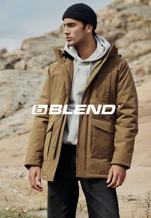 Bruine gewatteerde jas met capuchon, voorzijde met rits, twee grote zakken, gedragen over een grijze hoodie, in combinatie met donkere broek en een gebreide muts.