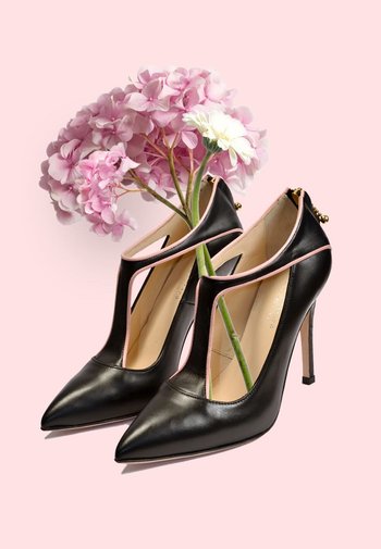 Zwarte leren hoge hakken met een spitse neus, open zijkanten, roze accenten en gouden hardware, gestyled met bloemen in de binnenkant van de schoen.