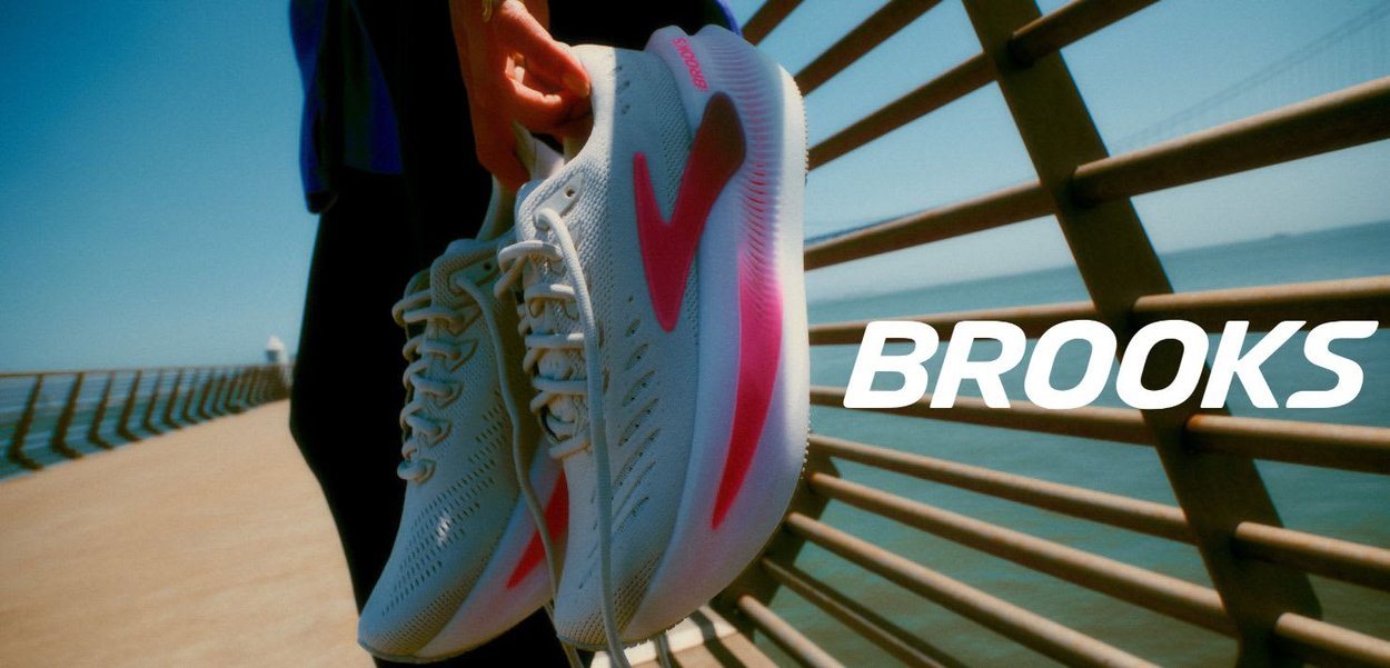 Witte hardloopschoenen met roze accenten, textuurrijk materiaal en een gestroomlijnd ontwerp, vastgehouden in een hand tegen een heldere buitenachtergrond.