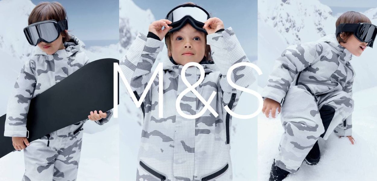 Dziecko w szarym kombinezonie zimowym w kamuflaż i goglach narciarskich, trzymające deskę snowboardową i poprawiające gogle w ośnieżonym górskim otoczeniu.