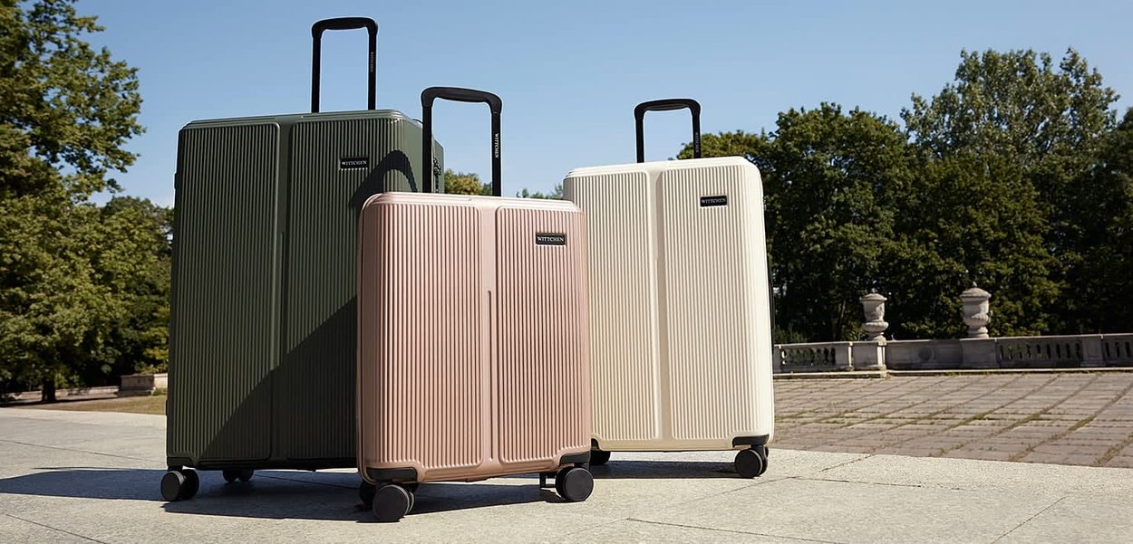 Trzy walizki w kolorze zielonym, różowego złota i kremowym z pionowym żebrowaniem, czarnymi uchwytami i kółkami, ułożone w linii na świeżym powietrzu.