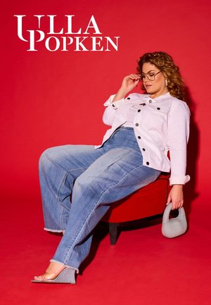 Witte denim jas met zwarte knopen, oversized lichtblauwe spijkerbroek, glazen met heldere montuur en grijze pantoffels tegen een helderrode achtergrond.