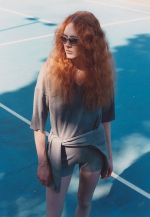 Top grigio, morbido e ampio sopra pantaloni corti abbinati, legato in vita. Indossato con occhiali da sole scuri. Ambientazione: superficie di un campo da tennis blu.
