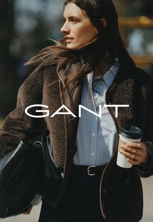 Bruine fleecjas over een blauw gestreept shirt, houdt een koffiekop vast, met een zwarte leren tas. Tekst: "GANT" prominent weergegeven.