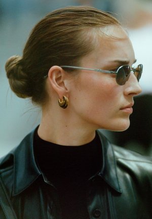 Veste en cuir noire, col roulé noir, lunettes de soleil à monture argentée et boucles d'oreilles dorées. Cheveux coiffés en un chignon bas, arrière-plan neutre.