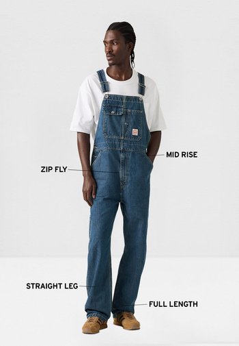 Hombre vestido con overol de mezclilla azul con cierre de cremallera, cintura de tiro medio, pierna recta y diseño de longitud completa, combinado con camiseta blanca y zapatos marrones.