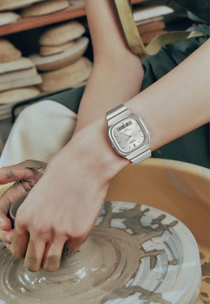 Manos moldeando arcilla en un torno de cerámica, con un reloj de pulsera Casio plateado, y estantes con herramientas y materiales de cerámica en el fondo.