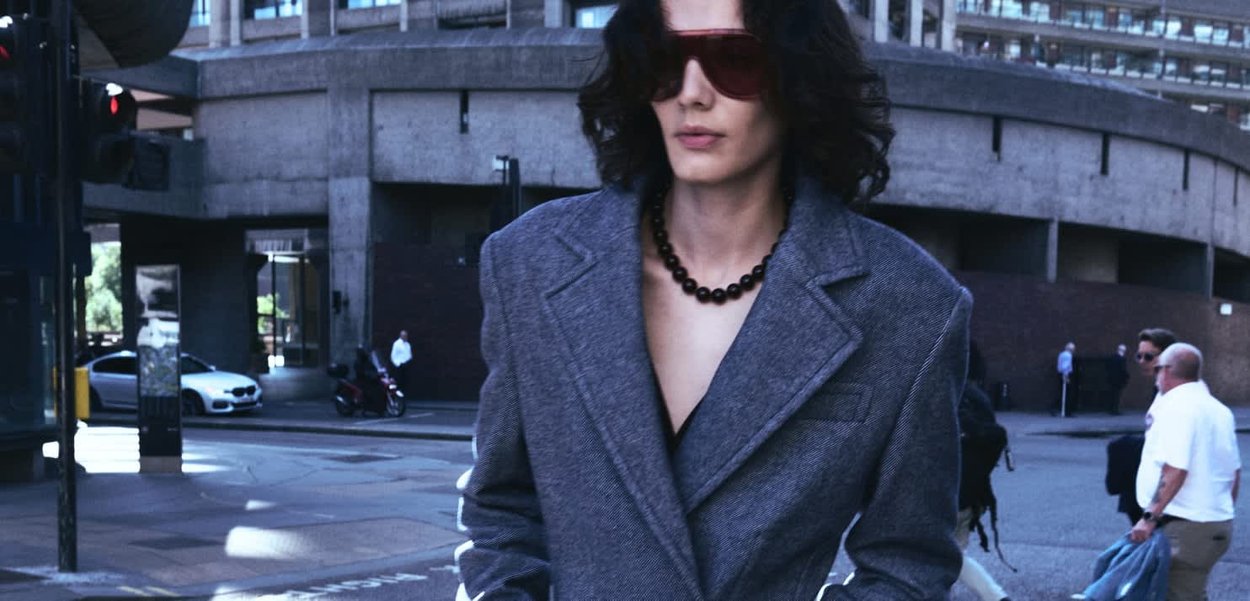 A gray oversized blazer with a textured finish, paired with red sunglasses and a black beaded necklace. Urban background with pedestrians.