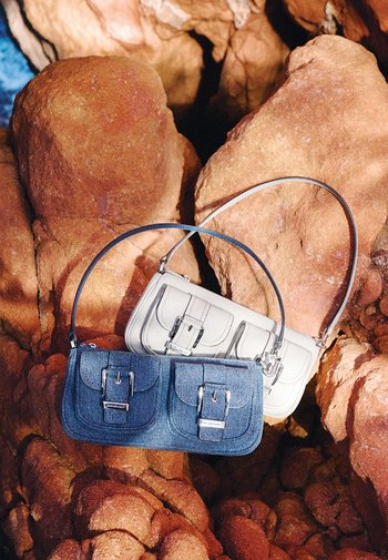 Two handbags on rock surface: one denim blue with dual front pockets and silver accents, one cream with a structured design and silver hardware.