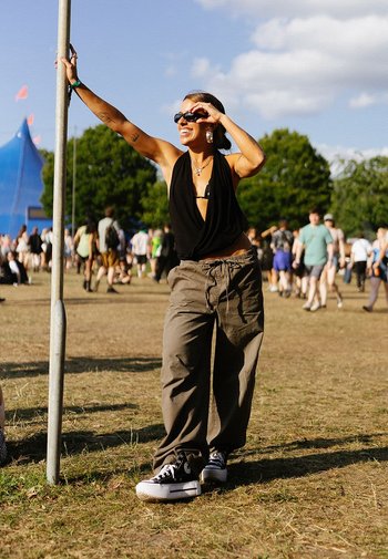 Black sleeveless top, olive green baggy pants, and black high-top sneakers. Standing by a pole at an outdoor event with a crowd behind.