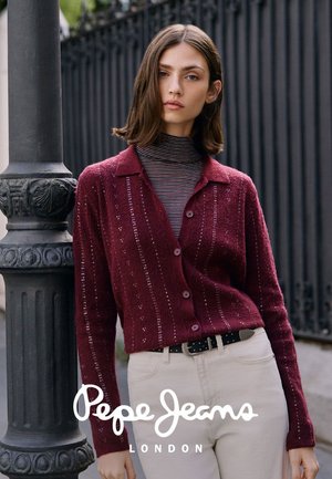 Mujer joven con un cárdigan marrón burdeos abotonado sobre un jersey de cuello alto a rayas y pantalones blancos, de pie junto a un farol negro decorativo.