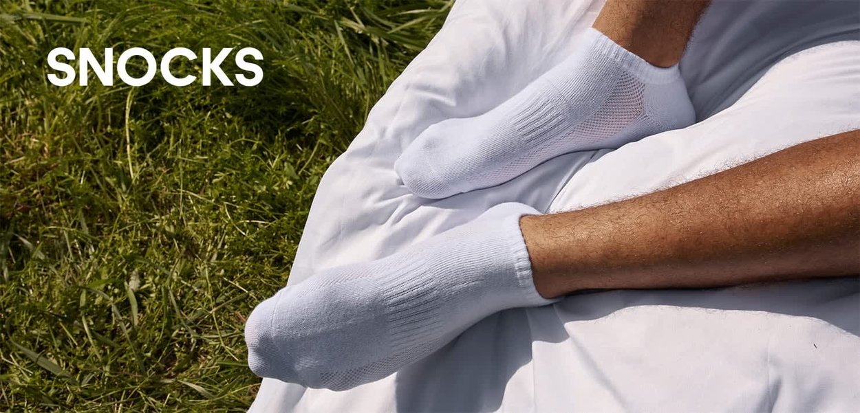 Zwei nackte Beine mit weissen Knöchelsocken ruhen auf einer weissen Decke auf grünem Gras mit dem Wort "SNOCKS" in weissem Text.