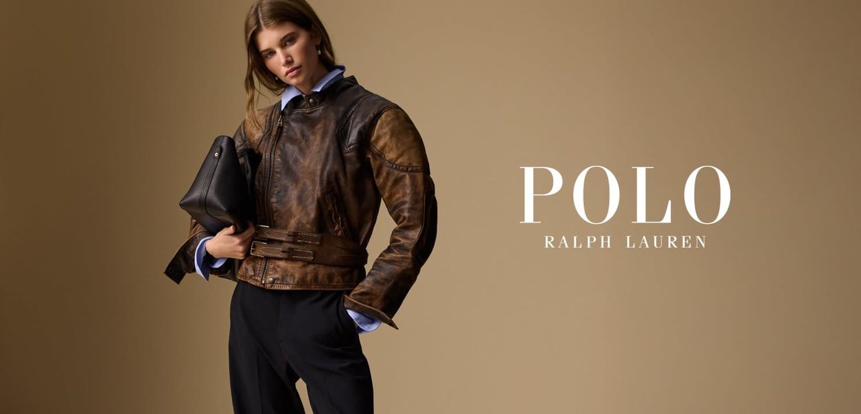 Chaqueta de cuero marrón con un diseño ajustado, cinturilla con hebilla y mangas largas; el modelo sostiene un bolso negro. Fondo liso, el texto incluye "POLO RALPH LAUREN."
