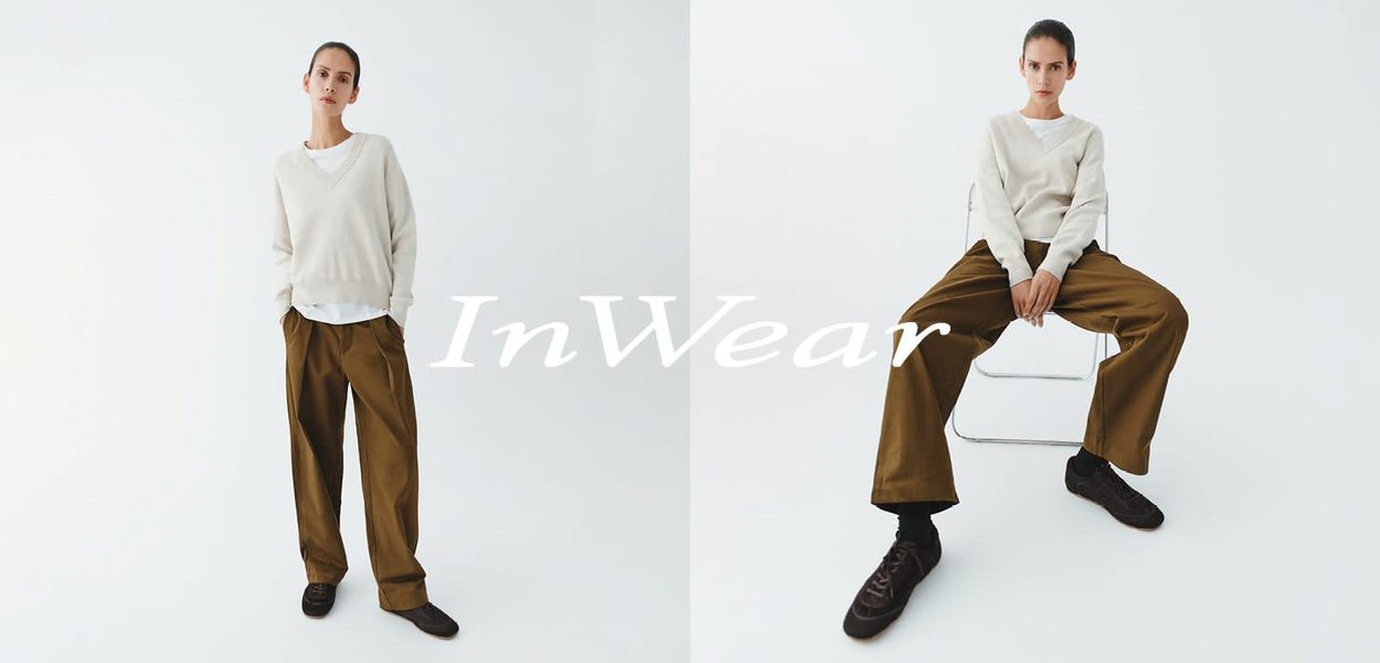 Beige V-Ausschnitt-Pullover, weiße Unterlage, braune Hosen mit weitem Bein und schwarze Turnschuhe; Model steht und sitzt auf einem transparenten Stuhl vor weißem Hintergrund.