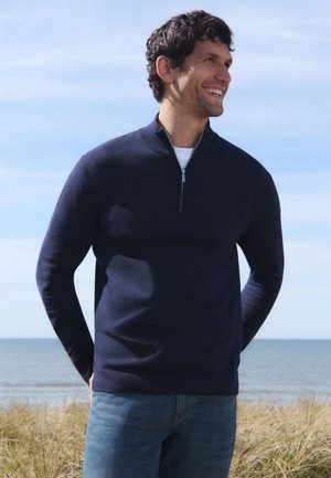 Ein Mann lächelt und schaut nach rechts, trägt einen navyblauen Halbzip-Pullover und Jeans. Er steht in der Nähe von Gras mit einem ruhigen Meer und Himmel im Hintergrund.