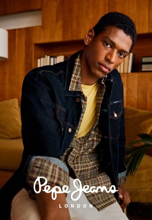 Joven vestido con chaqueta de mezclilla en capas, camisa de cuadros y camiseta amarilla, sentado en interiores con estanterías de madera y plantas al fondo.
