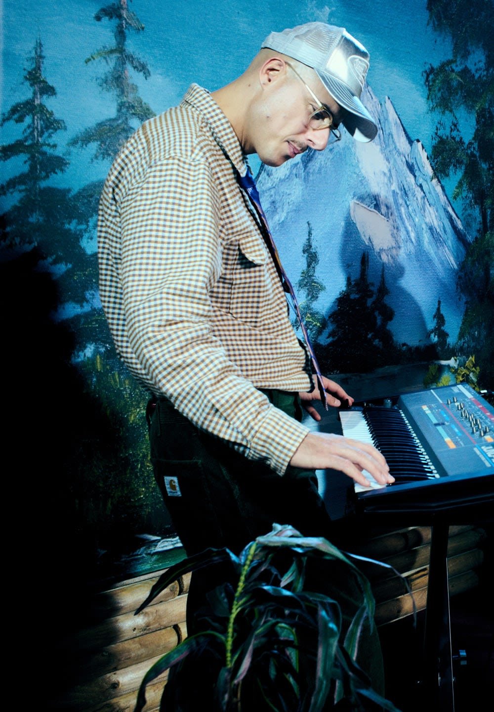 Elektroninis klaviatūras su juodomis klavišais ir spalvingais mygtukais. Asmuo su languota marškiniais ir žaliais džinsais groja. Fone matosi kalnų peizažas.