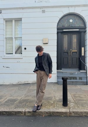 Zwarte leren jas, zwart shirt, beige broek en donkere schoenen. Staand op een stenen trottoir naast een zwarte paal voor een deur.
