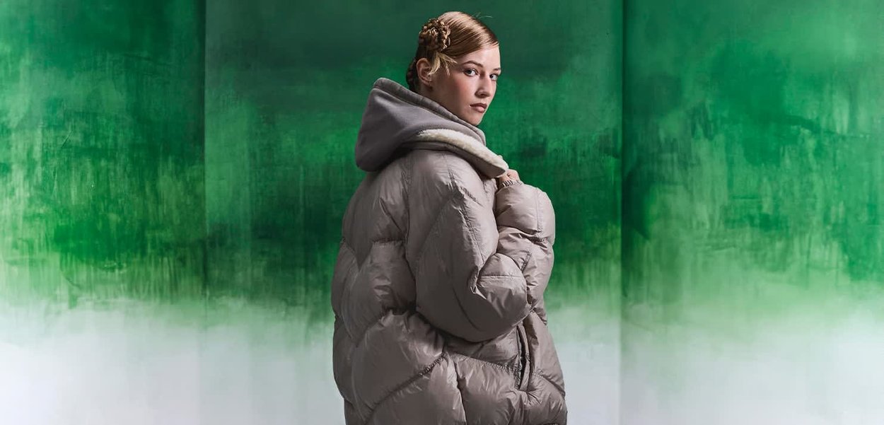 Doudoune de couleur gris clair, dotée d'une grande capuche et d'une finition texturée, avec un motif de matelassage en losanges. Le fond est en dégradé de vert.