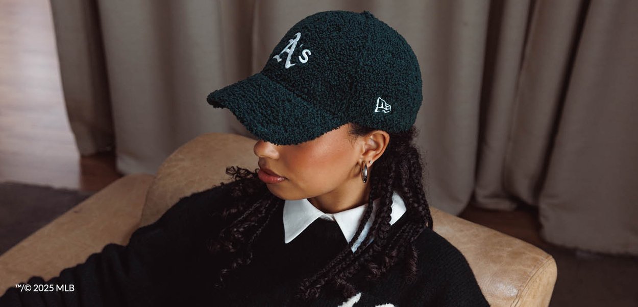 Donna con capelli intrecciati che indossa un cappellino da baseball nero con il logo "A's", maglione nero, camicia bianca con colletto e orecchini a cerchio, seduta al chiuso.