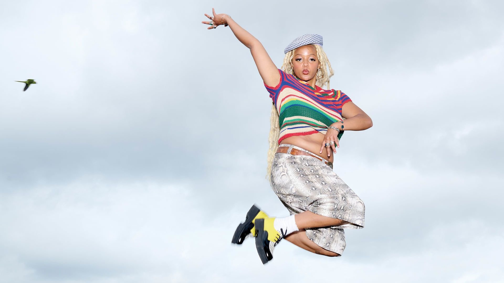 Kleurvolle gestreepte gebreide top, metallic slangpatroon rok en zwarte platform schoenen met gele accenten, tegen een bewolkte lucht.