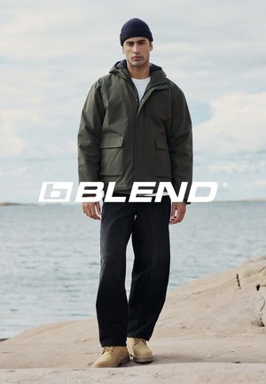 Veste imperméable vert olive avec une capuche, deux poches avant, portée sur une chemise blanche, associée à un pantalon noir et des chaussures jaunes.