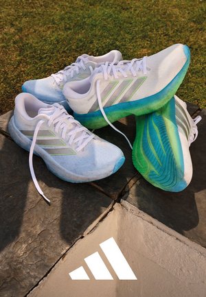 Par hvite og blågrønne joggesko med skolisser, plassert på steinfliser over gress, Adidas-logo synlig på bakken.