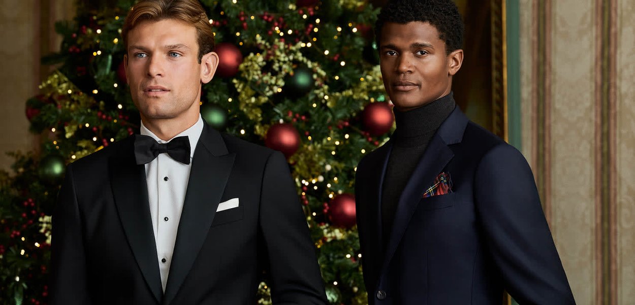 Dos hombres con ropa formal están frente a un árbol de Navidad decorado. Uno lleva un esmoquin negro con pajarita, el otro un traje azul marino con un cuello de tortuga.