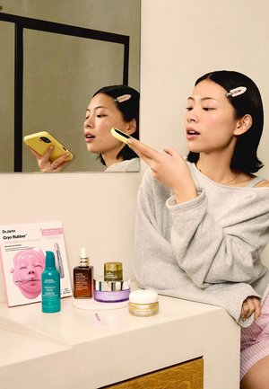 Vrouw met roze haarklem die in een gele smartphone spreekt terwijl ze zit aan een badkamermeubel met huidverzorgingsproducten.