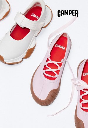 Dwie pary butów Camper z brązowymi podeszwami i czerwonymi wkładkami, jedna biała z paskiem na rzep, a druga różowa sznurowana, na białym tle.