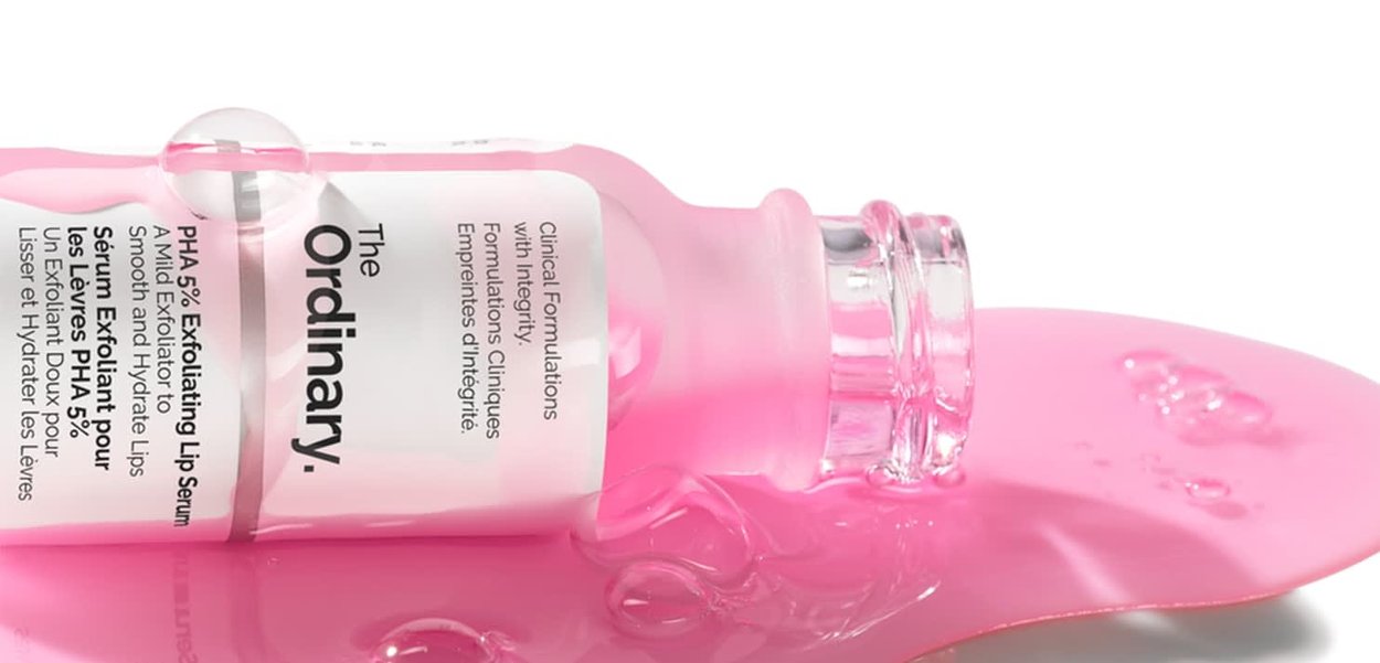 Clear glass bottle with a pink cap, containing a pink serum. A label displays product details. Serum spills on a glossy pink surface.