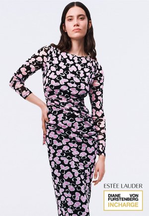 Donna con capelli lunghi e scuri che indossa un abito nero aderente con motivi floreali astratti rosa e bianchi, in piedi con una mano sul fianco.