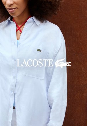 Woman wearing a light blue button-up shirt with a green crocodile logo on the chest pocket against a brown background.