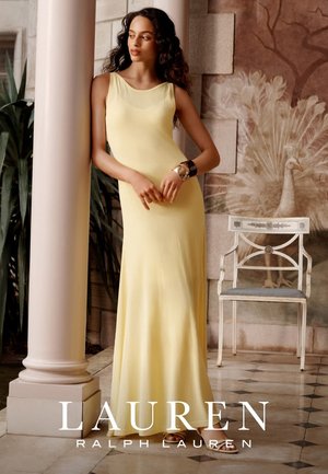Mujer con un vestido amarillo sin mangas y largo hasta el suelo apoyada en una columna con un mural decorativo de pavo real y una silla blanca al fondo.
