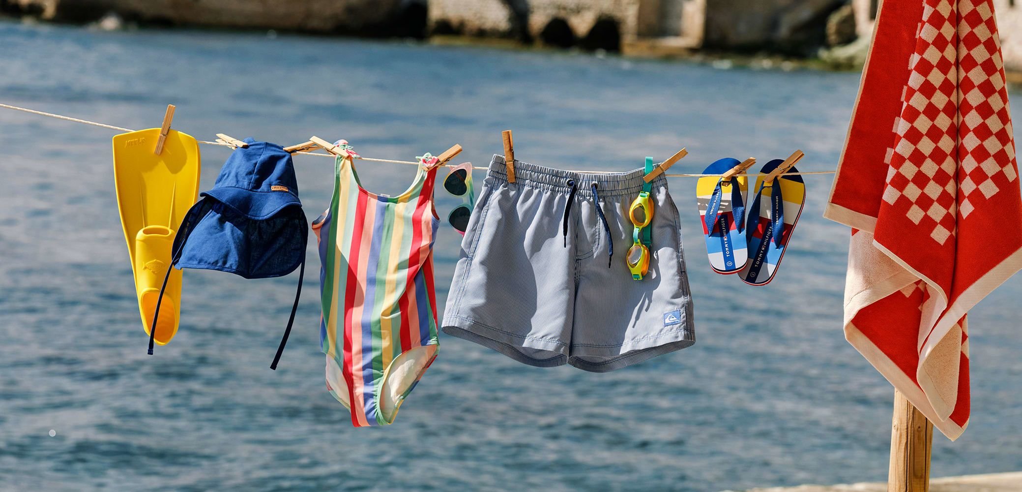 Plivanje oprema visi na konopcu: žute peraje, plava kapa, šareno prugasti kupaćic, sivi šorts, žute sunčane naočale, plave japanke, crveni ručnik.