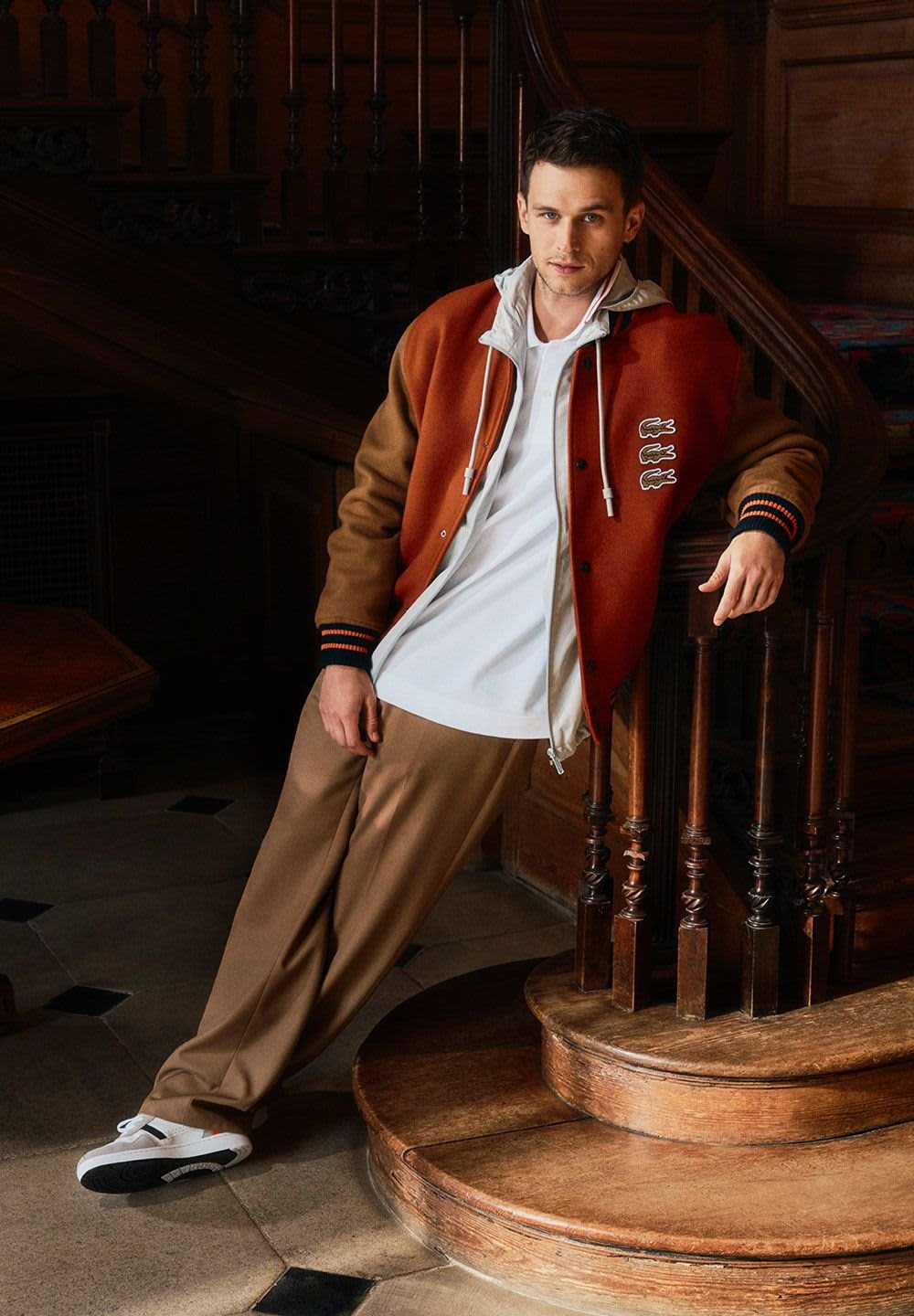 Veste zippée marron et orange superposée sur un t-shirt blanc, associée à un pantalon beige et des baskets blanches ; debout à côté d'un balcon en bois.