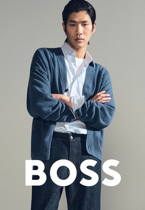 Junger Mann mit verschränkten Armen, der eine blaue Strickjacke, ein gestreiftes Hemd und dunkle Jeans trägt, vor einem schlichten Hintergrund mit dem Schriftzug "BOSS" steht.