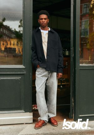 Dark button-up jacket over a light sweater, grey loose-fitting jeans, and brown shoes. Standing at an open door with a green frame.