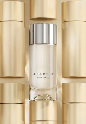 Flacon de parfum Le Sel d'Issey avec un corps en verre transparent, bouchon métallique argenté, entouré de contenants cylindriques dorés. Texture lisse.
