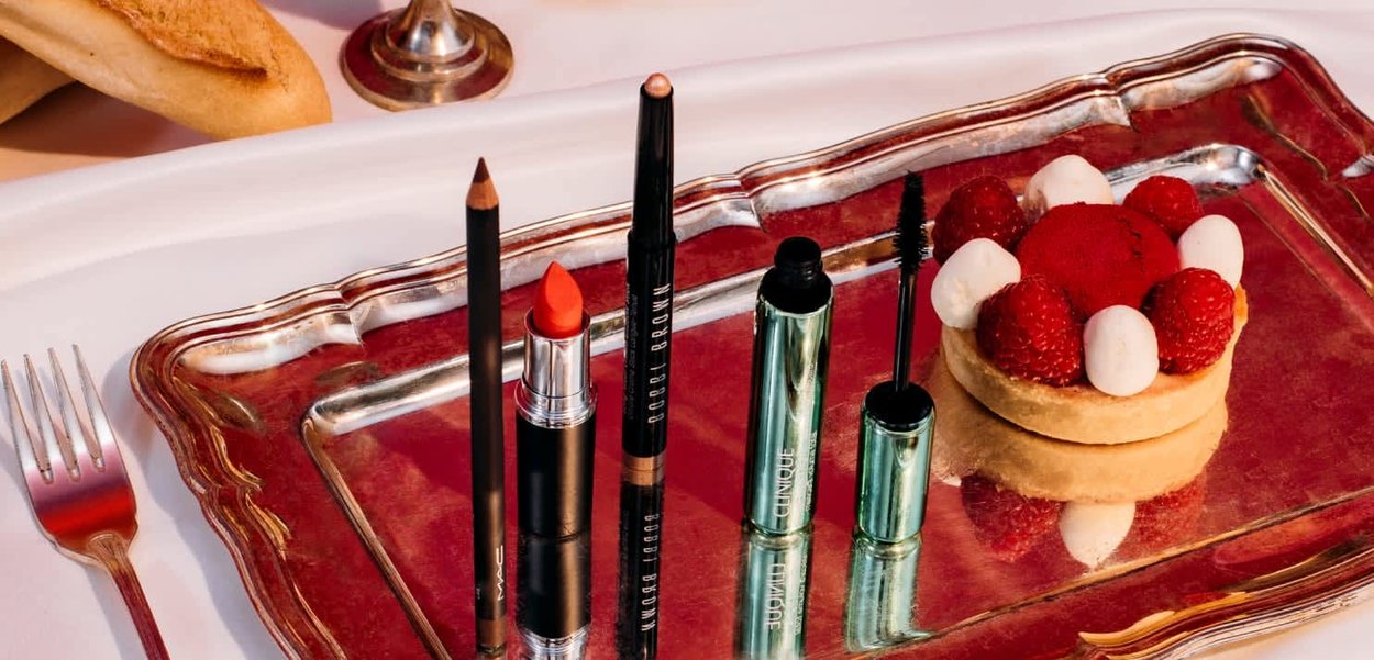Sei prodotti di trucco su un vassoio d'argento, tra cui eyeliner nero, rossetto arancione, mascara e un dolce con lamponi su una crostata.