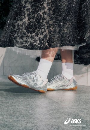 Legs wearing white socks and silver Asics sneakers, partially covered by a black floral lace skirt, standing on gray floor tiles.