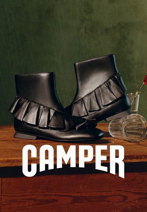 Paar schwarze Leder-Stiefeletten mit Rüschendetails, auf einem Holztisch neben einem umgekippten Glas und einer kleinen Blumenvase platziert.