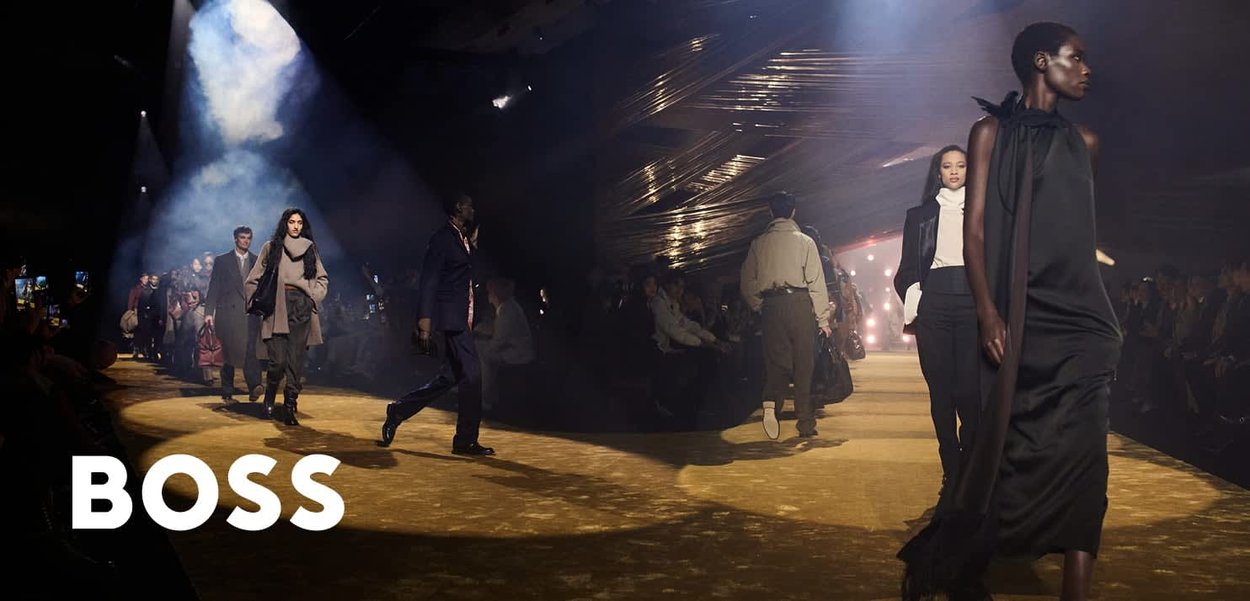 Modelki chodzą po lekko oświetlonym wybiegu, prezentując stylowe, różnorodne jesienne stroje podczas pokazu mody BOSS z dramatycznym oświetleniem.