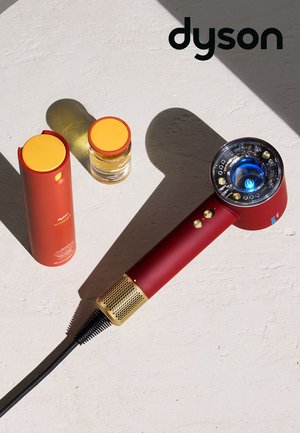 Asciugacapelli Dyson rosso con dettagli dorati, cavo collegato, flaconi per la cura dei capelli con tappo giallo e logo Dyson su una superficie testurizzata.