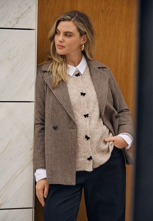 Bruine houndstooth blazer over een beige gebreide trui met zwarte knopen, gecombineerd met donkere broeken. Opvallende gouden creolen.