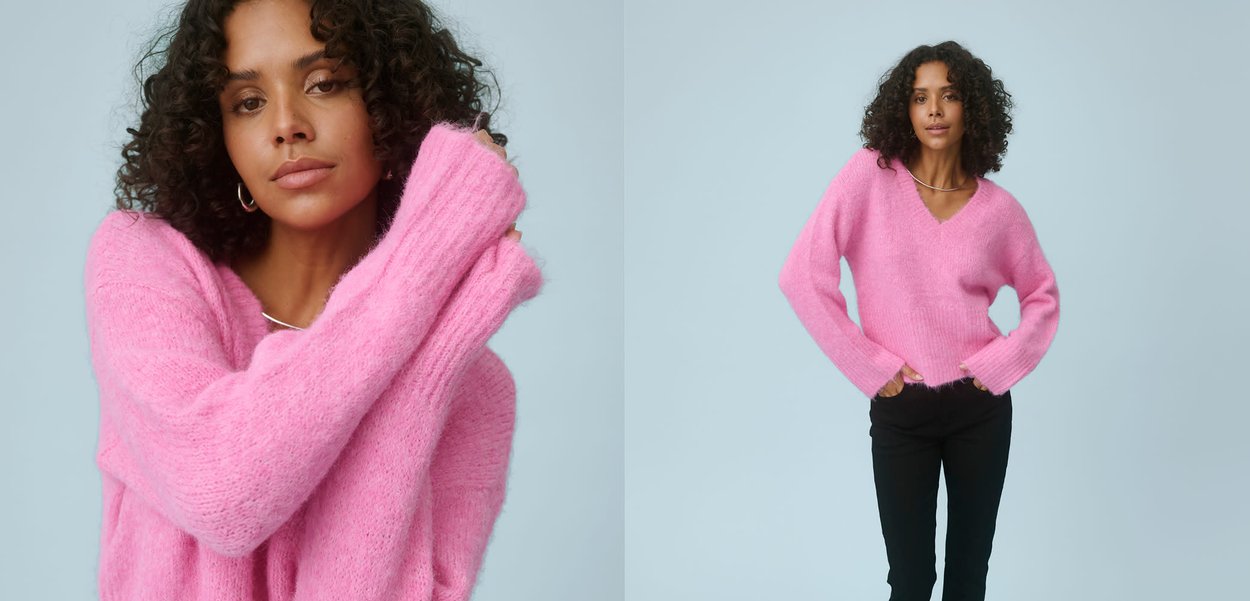 Frau mit lockigem Haar, die einen zartrosa Pullover trägt und mit einem erhobenen Arm vor einem einfarbigen hellblauen Hintergrund posiert.